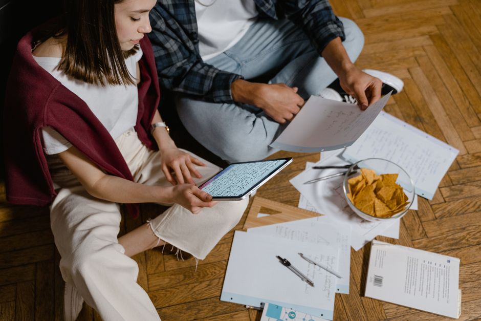 A man and woman study together on a wooden floor using technology and papers